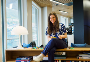 A brown haired woman smiling as she sits on top of a low book shelf.