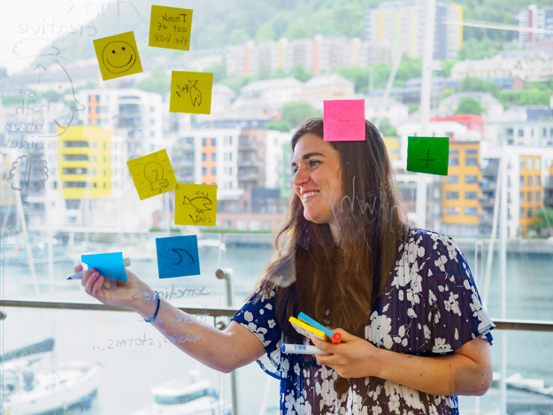 A woman seen from the other side of a glass window. Surronded by colourful post-its as she hangs another one up.