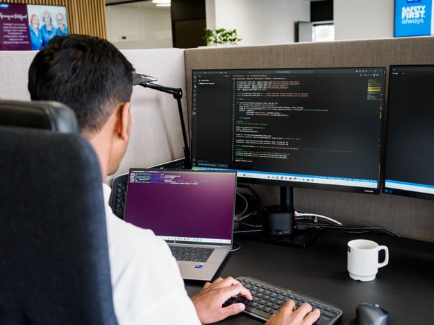 Over the shoulder picture of a man sitting in front of a computer. On his computer is a screen full of codes. 